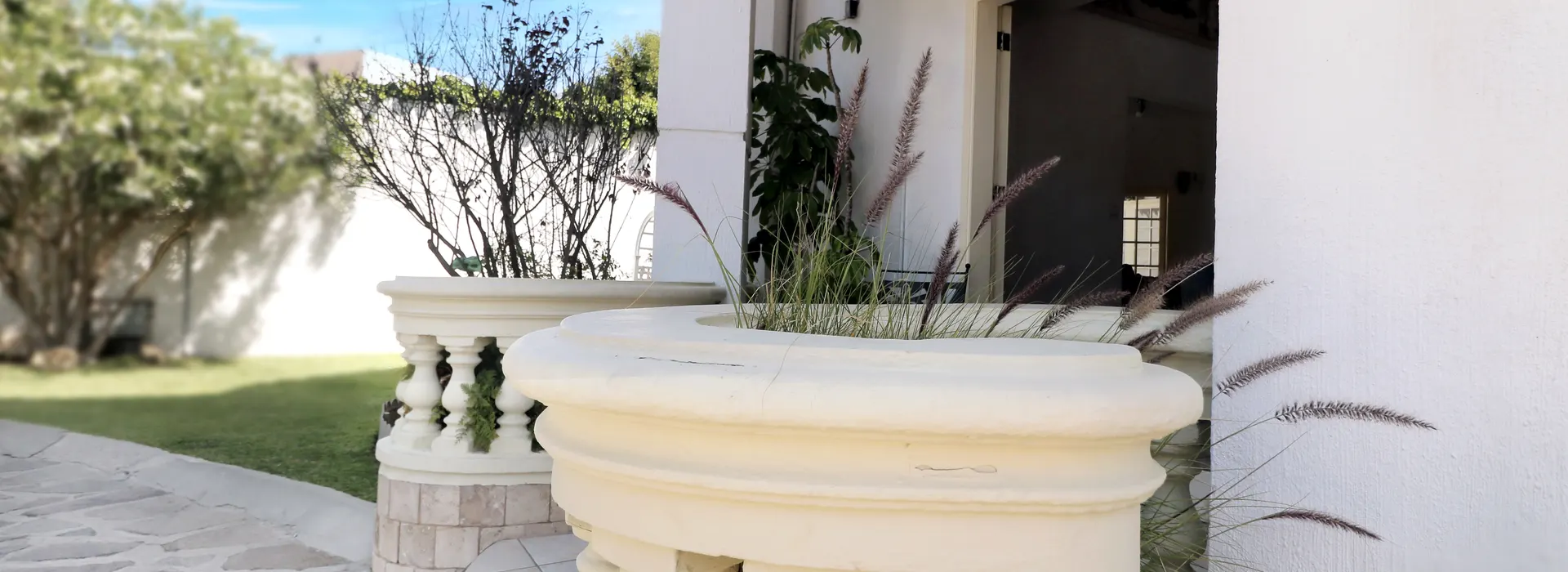 A white balustrade with ornamental grasses frames an outdoor patio area. Potted plants and trees stand in the background under a partly cloudy sky.