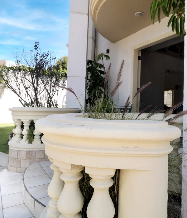 A white balustrade with ornamental grasses frames an outdoor patio area. Potted plants and trees stand in the background under a partly cloudy sky.