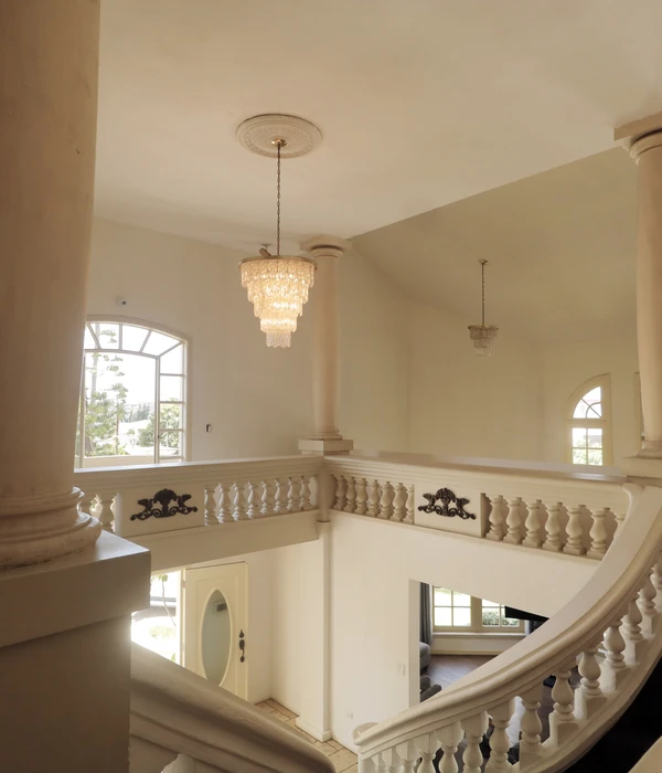 Bright interior of a building featuring a grand staircase with stone railings, pillars, and elegant chandeliers.