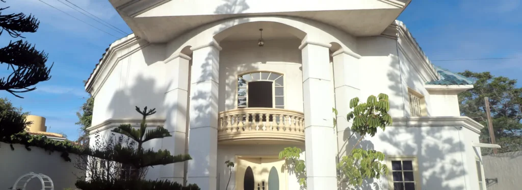 Large white building entrance with tall columns, a curved balcony, and double doors under a bright blue sky.