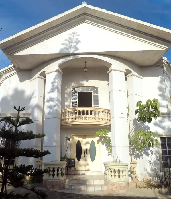 Large white building entrance with tall columns, a curved balcony, and double doors under a bright blue sky.