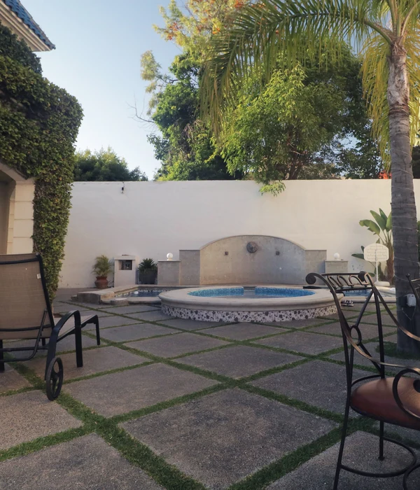A patio with stone tiles and grass borders, featuring a circular pool, a fountain wall, palm trees, and lounge chairs.