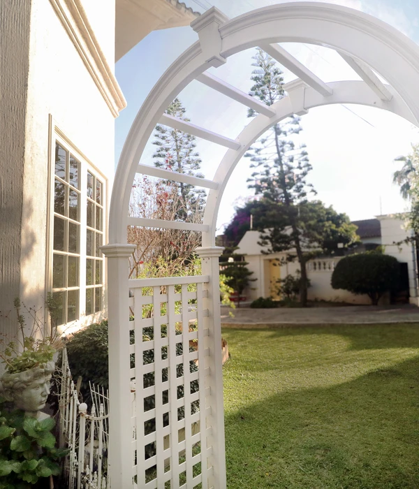 White wooden garden arbor with a lattice side panel on a green lawn, next to a sunlit building and tall pine trees.