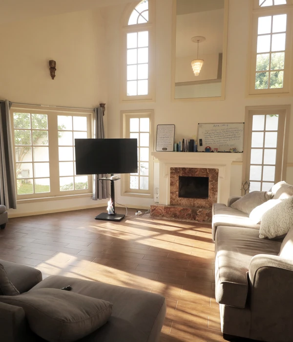 Sunlit common room with wood-patterned floor, grey sectional, TV on a stand, marble fireplace, and tall windows.