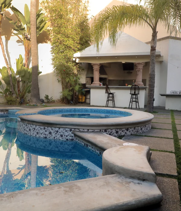 Outdoor pool and built-in circular jacuzzi with tiled edge, next to a paved patio with palm trees and a covered bar area.