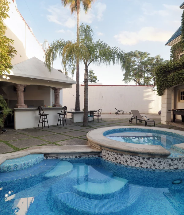 Outdoor swimming pool and circular jacuzzi in a stone patio with palm trees, lounge chairs, and a covered outdoor bar area.