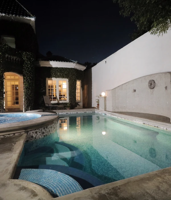 Outdoor pool and jacuzzi at night, featuring a stone wall fountain, lit windows, and ivy-covered building walls.